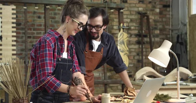 Easy Communication in Designer Studio/Two hipsters having a small talk during their work in a wood workshop. They are very positive and happy, smiling and making jokes. The man looks at the girls work