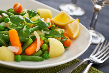 Salad with baby carrots, peas, onions and potatoes served on white plate on black wooden background