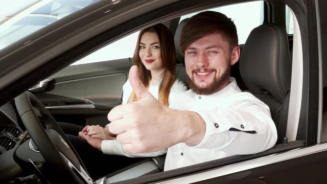 Handsome bearded guy showing his thumb up through the car window. Close up of young caucasian man approving new car at the dealership. Nice couple smiling for the camera from inside the car