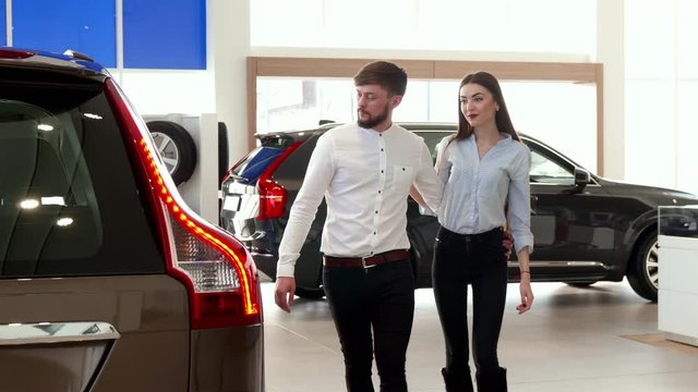 Young couple walking at the car dealership. Handsome brunette guy and his girlfriend looking at the one of cars at the showroom. Caucasian bearded man and brown haired woman standing against