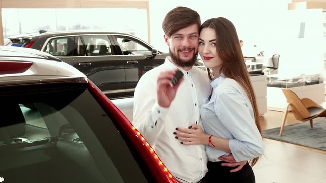 Nice couple embracing each other near the car at the dealership. Brunette bearded man and his girlfriend look at each other againts background of black SUV. Young caucasian guy holding car key in his
