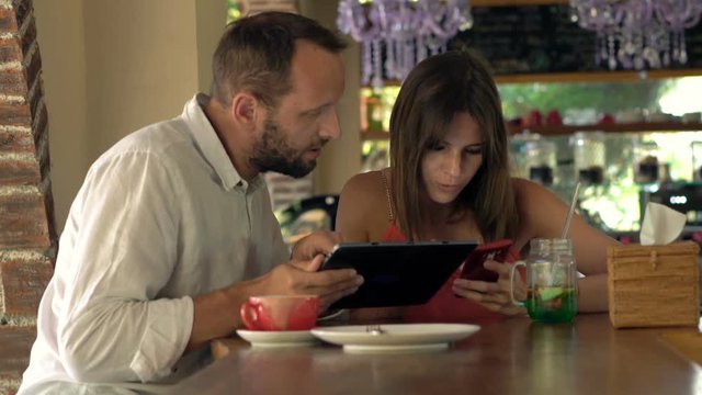 Young couple with tablet and smartphone talking sitting by counter in cafe
