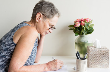 Profile view of older woman with short grey hair and glasses sitting at desk writing (selective focus)
