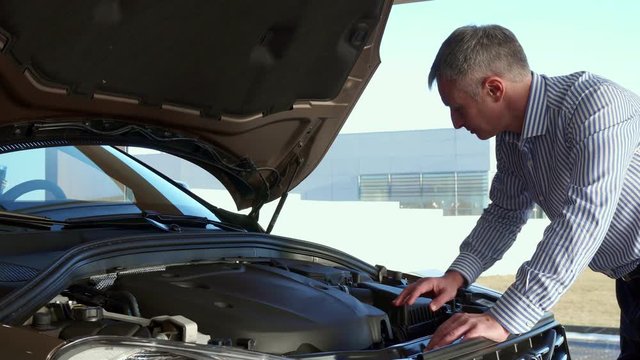 Attractive mature man looking at the car engine at the dealeship. Caucasian gray customer examining engine compartment of the car. Attractive middle aged client moving away from the vehicle