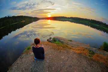 Young woman sitting on the stone enjoying peaceful moment of sunset. In the reflection of the lake...