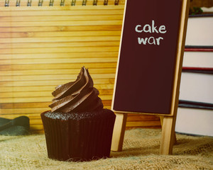 Delicious Muffin by the board that says CAKE WAR, background set of books on sackcloth