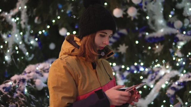 Beautiful young woman in bright winter clothes is messaging on the smart-phone at the christmas tree background in the evening.