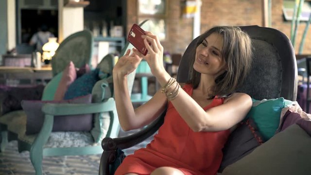 Young, pretty woman taking selfie photo with cellphone sitting on sofa in trendy cafe
