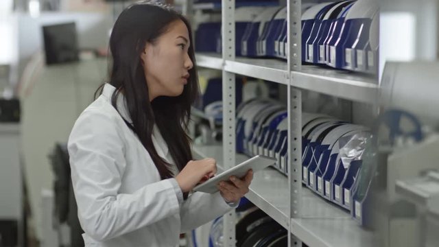 Tracking shot of young Asian woman working on digital tablet while inspecting shelves in laboratory