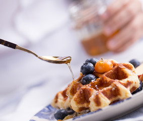 Blueberry apple waffle with honey set on a dish with a chef's hand holding spoon for a jar of honey ready to serve.