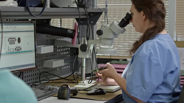 Tracking shot of women examining printed circuit board with digital microscope connected with computer monitor