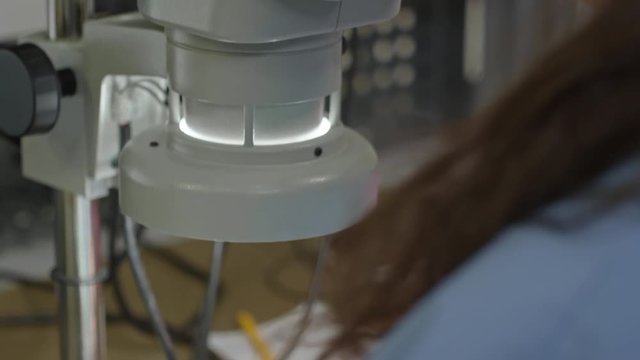 Tilt down of female technician examining PCBA through microscope in laboratory
