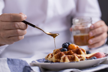 Blueberry apple waffle with honey set on a dish with a chef's hand holding spoon for a jar of honey ready to serve.