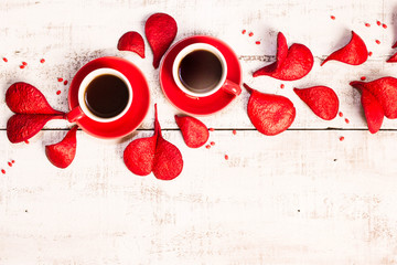 Two red coffee cups, petals in the shape of hearts on a white wooden background