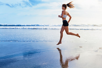 Active sporty woman run along ocean surf by water pool to keep fit and health. Sunset black sand beach background with sun. Woman fitness, morning workout and sport activity on summer family holiday.