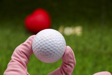 hand of lady golfer is holding golf ball with blurry red heart shape on green grass background. 