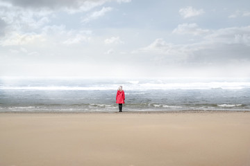 single woman in red raincoat standing on a beach looking at the stormy sea