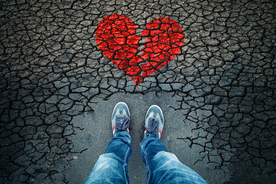 Lonely man standing on cracked asphalt floor with illustrated cracked broken heart symbol. Point of view perspective used.