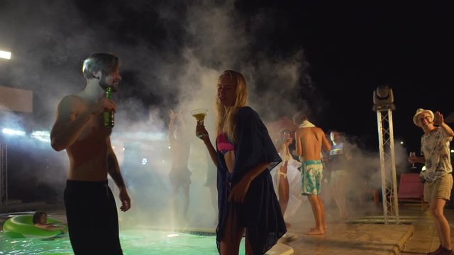 Topless Caucasian male with dark beard and beer dancing with blonde female with cocktail at night pool party with people on background. In slowmotion