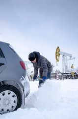Man shoveling snow to free stuck car. Winter period. Industrial site and pump jack background. 