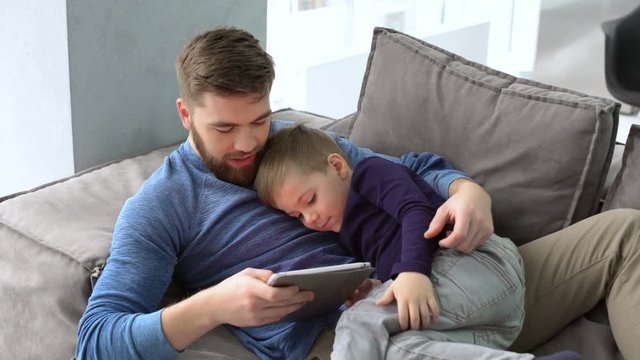 Father and his young son surfing on a tablet while laying on a sofa ay home 