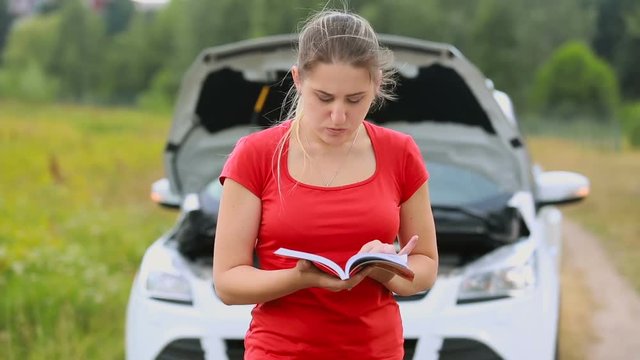 Upset young woman reading standing at brokedown car and reading manul on the deserted countryside road