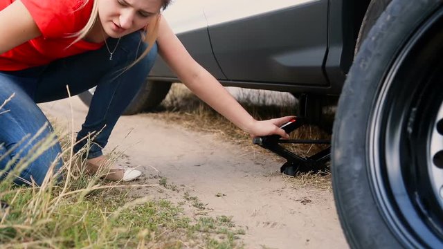 Young woman using jack-screw to lift a car before changing flat tire on dirt road at countryside