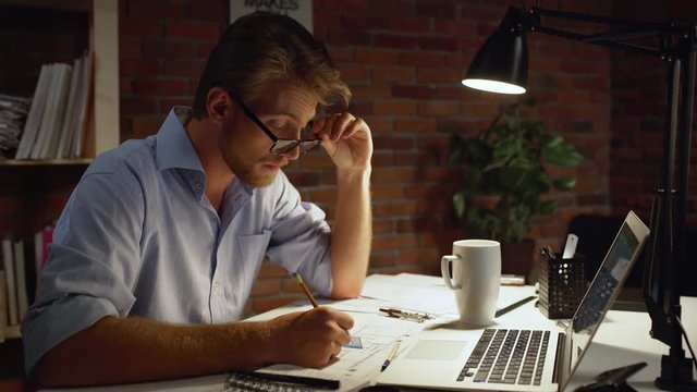 Young Caucasian designer concentrated taking off glasses drwing with pencil sitting at table near computer.