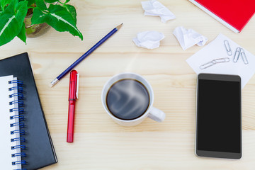 Hot coffee cup with smartphone, crumpled paper, glasses, notebooks, pen, pencil, paper clips, green leaves pot on vintage wooden table