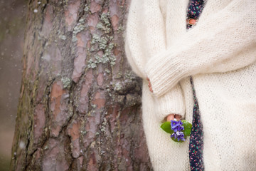 Young woman in white cardigan standing near an old tree and  holding beautiful snowdrops in her hands. First spring flowers in a forest. Beginning of spring in a forest.
