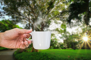 Hand holding hot coffee cup in morning on blurred park with sunlight and flare background