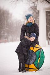 Fototapeta premium Young smiling woman and cute kid boy with inflatable sled walking in a winter park. Happy family enjoying snow walk. Mother and son. Lifestyle concept