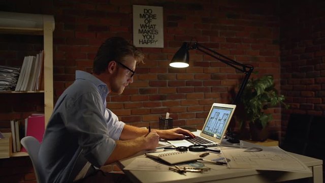 Young successful acrchitect with light beard and glasses sitting drawing with stylus at computer writing notes with table lamp. In slowmotion