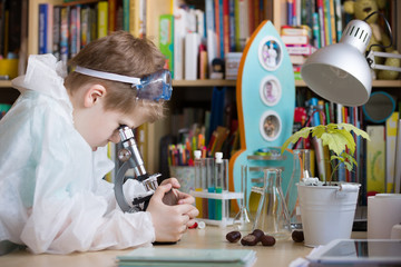 Cute elementary school boy looking into microscope at his desk at home. Young scientist making experiments in his home laboratory. Indoors. Child and science.