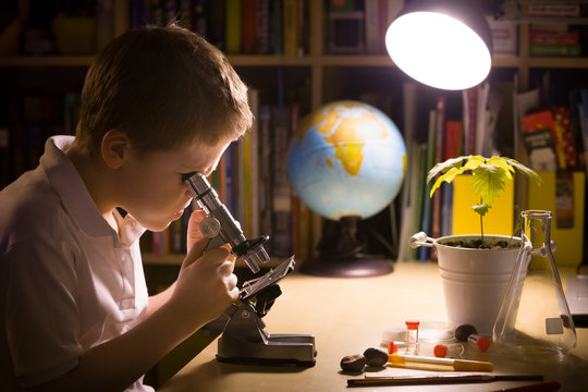 Close-up portrait of young student working with microscope in his room. Child and science experiments. Kid studying samples under the microscope. Preparing for science lesson.
