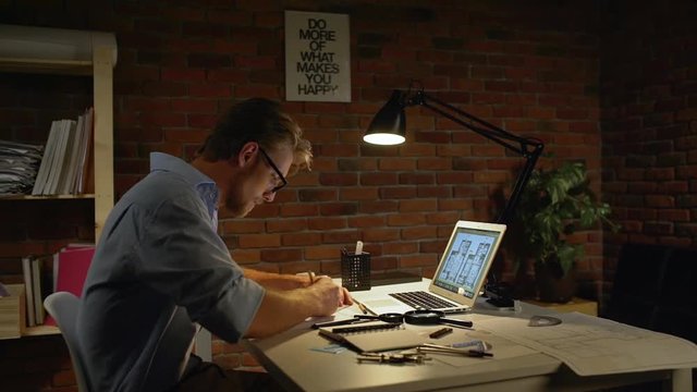 Young concentrated acrchitect with light beard and glasses sitting drawing with pencil by computer writing notes with table lamp in slowmotion