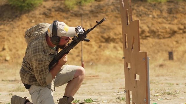 Bearded Caucasian male with headphones and sunglasses reloading and firing airsoft gun behind cover position in slowmotion outdoors.