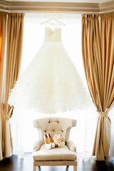 Gorgeous wedding dress hangs on the window over soft chair with