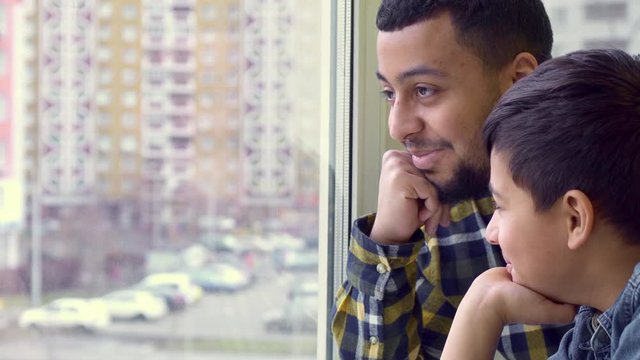 Attractive Father And Son Looking Out The Window. Close Up Of Little Brunette Boy And His Parent Leaning Chins On Their Hands. Side View Of Young African American Man And His Child Enjoying City