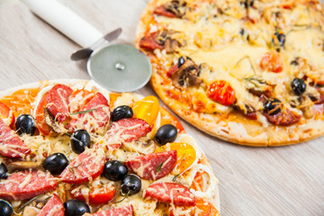 Pizza cutter together with two large pizzas with cheese olives and salami, mushrooms and tomatoes. On a wooden stand