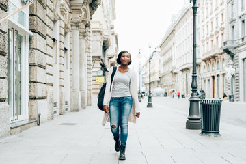 Young beautiful black woman outdoor in the city, looking at camera smiling wearing back pack - happiness, carefree, serene concept