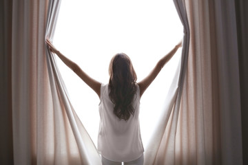 Beautiful woman opening curtains and looking through the window