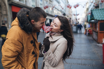 Fototapeta premium Beautiful young couple in love enjoying together at city street and eating together a lollipop in heart shape.