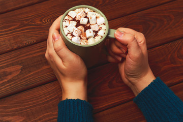 Hot cocoa or coffee mug and marshmallows with cinnamon in female hands on wood 