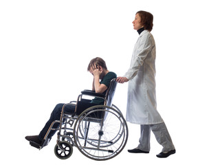 Young distressed boy sitting in a wheelchair holding his head and behind a wheelchair stands a woman in the medical gown on a white background