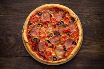 Homemade italian pizza with ham, tomatoes, olives, cheese on dark rustic wooden background.