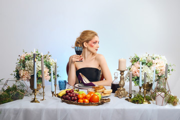 Fashion blonde model with colorful makeup drinking wine behind decorated table and eating suede shoe