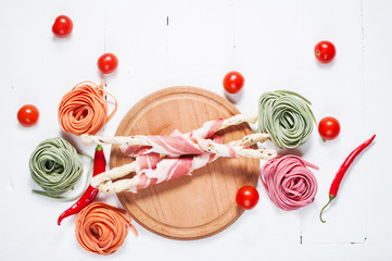 Colorful rolled pasta, cherry tomatoes and bacon on the white wooden background