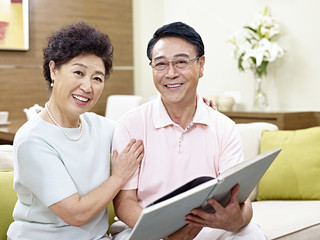 senior asian couple sitting on couch holding a book looking at camera