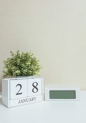 White wooden calendar with black 28 january word with clock and plant on white wood desk and cream wallpaper textured background , selective focus at the calendar